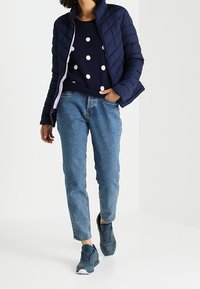 Granatowa kurtka puchowa o pikowanej teksturze, biały sweter w kropki, jasnoniebieskie jeansy i szare trampki. Casualowy zestaw na chłodną pogodę.