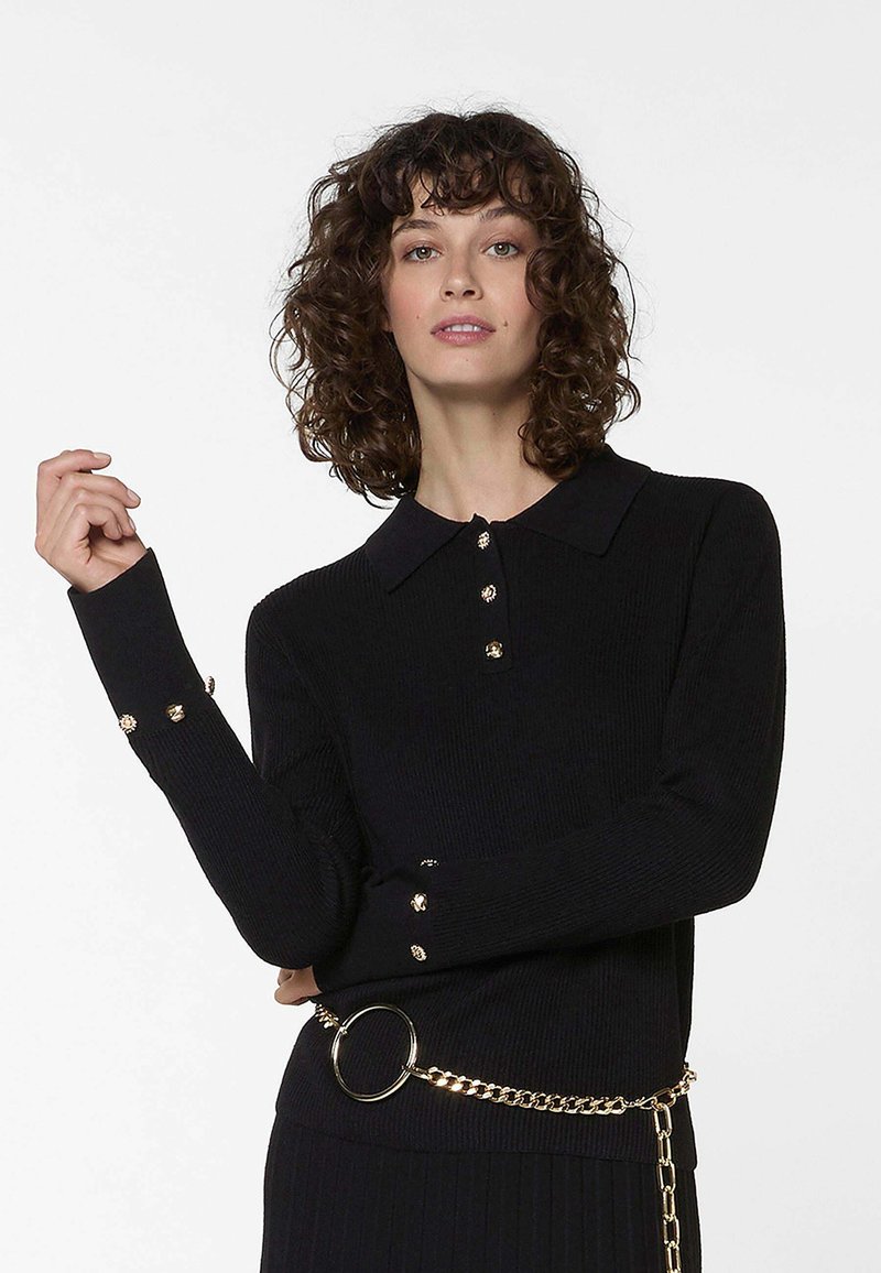 Vrouw met krullend haar draagt een zwarte geribbelde trui met gouden knopen en een gouden kettingriem tegen een effen witte achtergrond.