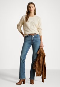 Pull en tricot crème avec logo en relief, jean flare en denim bleu, ceinture en cuir brun, et veste en cuir brun tenue à la main, bottes à talons brunes.