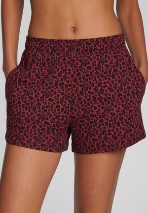 Frau trägt hoch taillierte bordeauxrote Shorts mit schwarzem Leopardenmuster und elastischem Bund, Hände in den Seitentaschen.