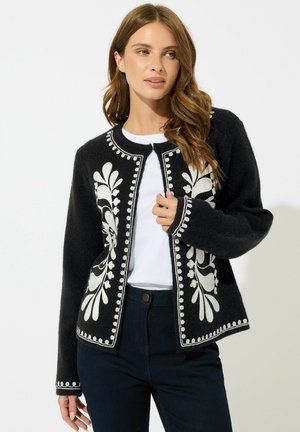 Donna che indossa un cardigan nero con ricami floreali bianchi sopra una camicia bianca e pantaloni scuri, posando davanti a uno sfondo chiaro e uniforme.