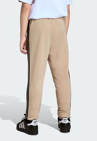 Beige track pants with black side stripes, featuring a tapered design and smooth fabric. Paired with black shoes displaying three white stripes.