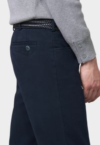 Navy-Hosen aus glattem Stoff mit subtiler Textur. Ein geflochtener schwarzer Gürtel sorgt für einen Kontrast, während ein dekoratives Detail an der Gesäßtasche hinzugefügt wurde.