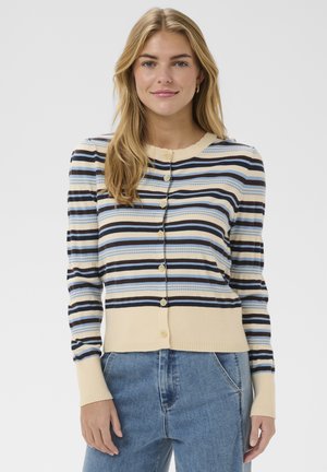 Mujer rubia con un cárdigan de rayas abotonado en colores beige, azul, blanco y negro, combinado con unos vaqueros azules, posando frente a un fondo liso.