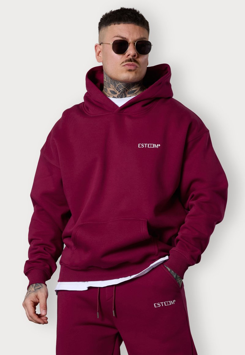 Sweatshirt à capuche bourgogne en tissu doux, avec une poche kangourou et un logo brodé. Le modèle porte des lunettes de soleil rondes et un pantalon de jogging assorti.