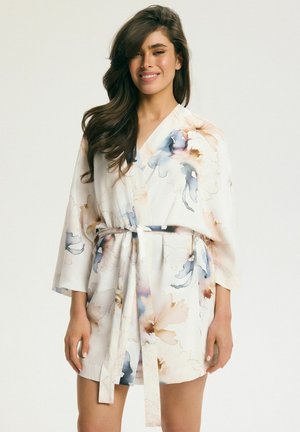 Kobieta uśmiechająca się, ubrana w biały kwiatowy szlafrok z niebieskimi i beżowymi kwiatami w stylu akwareli, związanego w talii paskiem.