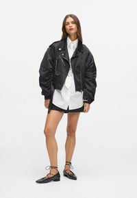 Giacca bomber in raso nero con vestibilità oversize, cerniere argento, indossata sopra una camicia bianca e una minigonna nera. Ballerine nere con borchie e dettaglio allacciato.