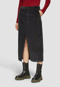 Jupe midi en velours côtelé noir avec une fente avant, fermeture à boutons et poches latérales, associée à des bottes chevilles noires et vertes épaisses.