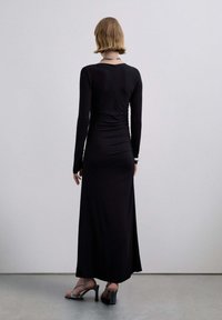 Vestido negro de manga larga con silueta entallada, detalle fruncido en la parte trasera y tela suave y elástica. Combinado con sandalias plateadas de tacón alto.