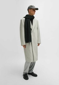 Cappotto di lana grigio chiaro, sciarpa nera con motivo geometrico, pantaloni grigi e scarpe slip-on nere, completati da un berretto grigio.
