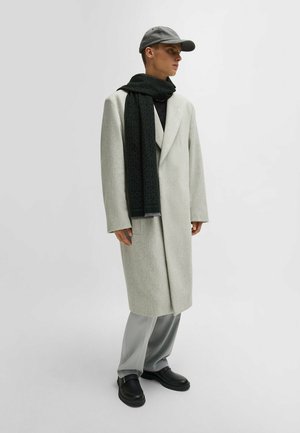 Light gray wool coat, black scarf with geometric pattern, gray trousers, and black slip-on shoes, complemented by a gray cap.