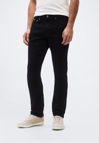 Μαύρο τζιν με στενή γραμμή, σχέδιο με πέντε τσέπες, ασημένια μεταλλικά εξαρτήματα και απαλό υλικό, συνδυασμένο με ανοιχτό μπεζ αθλητικά παπούτσια.