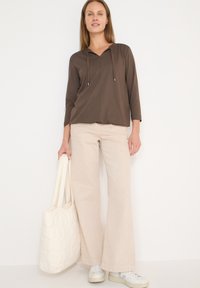Haut marron à capuche avec cordons, pantalon large beige clair, et sac cabas matelassé blanc, porté avec des baskets blanches.