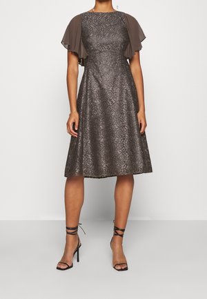 Kobieta w metalicznej koronkowej sukience do kolan z falbanowymi rękawami i czarnych sznurowanych sandałach na wysokim obcasie, stojąca na jednolitym tle.