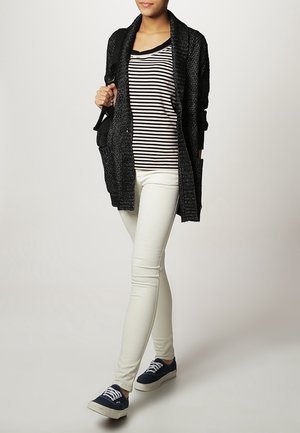 Donna che indossa un cardigan nero, una maglia a righe bianca e nera, pantaloni bianchi, sneakers blu navy e porta una borsa a tracolla nera, in piedi su uno sfondo semplice.