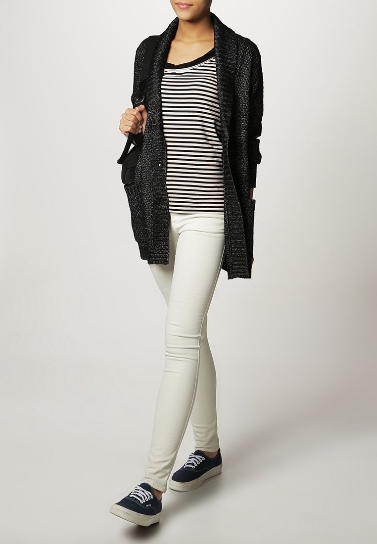 Femme portant un cardigan noir, un chemisier rayé noir et blanc, un pantalon blanc, des baskets bleu marine, et portant un sac à épaule noir, debout sur un fond uni.