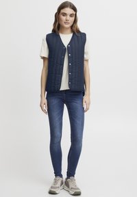 Marinblå quiltad väst med V-halsringning och tryckknappar, bärs över en ljusbeige skjorta. Kombinationen med mörkblå slim-fit jeans och grå sneakers.