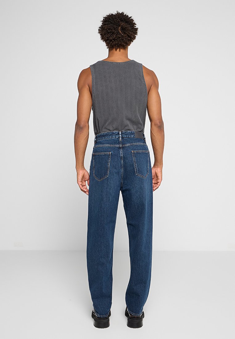 Mann in einem grauen ärmellosen Oberteil und hochgeschnittenen blauen Jeans, der mit dem Rücken zu einer schlichten hellen Hintergrund steht.