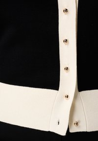 Zwarte cardigan met crèmekleurige accenten, voorzien van een knoopsluiting met gouden knopen en een geribbelde textuur aan de zoom en manchetten.