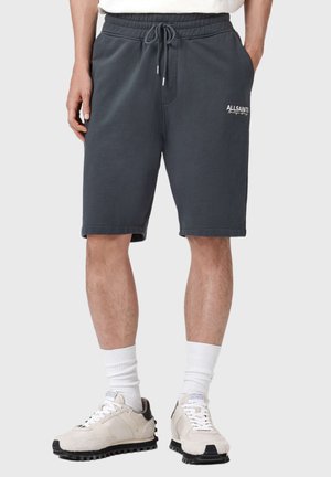 Mand iført mørkegrå AllSaints shorts med snører, hvide sokker og beige sneakers, stående med hænderne i lommerne mod en ensfarvet baggrund.