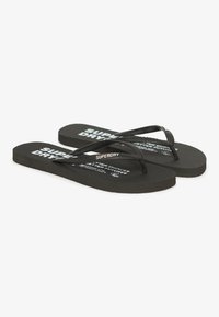 Tongs noires avec des semelles en caoutchouc texturé et des sangles. Présentent un texte de marque en blanc sur la semelle et la sangle.