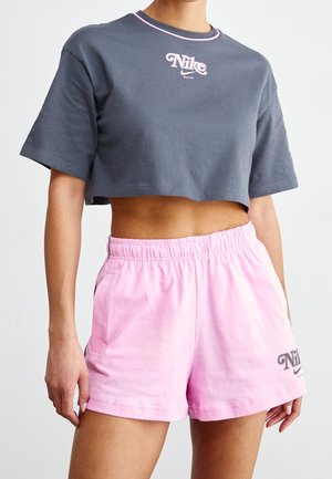 Frau trägt ein dunkelgraues, kurz geschnittenes Nike-T-Shirt und hellrosa Nike-Shorts mit elastischem Bund, posiert vor einem schlichten hellen Hintergrund.