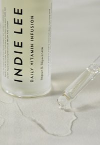Heldere glazen fles met het label "Indie Lee Daily Vitamin Infusion," met een druppelaar die een vloeistof op een gestructureerd oppervlak laat vallen.