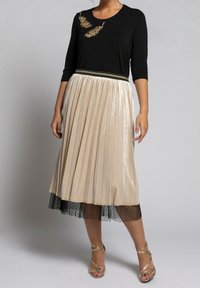 Haut noir avec broderie de feuilles, jupe plissée beige métallique superposée à un tulle à pois noirs, complétée par des sandales dorées à lanières.