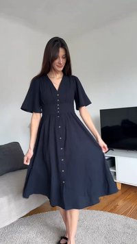 Žena v černých midi šatech s knoflíky vpředu a nafouknutými rukávy stojí v obývacím pokoji s pohovkou a televizí v pozadí.