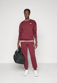Bordeaux sweater med en frontlomme og logo, parret med matchende joggers. Modellen holder en sort træningstaske. Hvide sneakers fuldender looket.