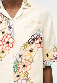 Λευκό πουκάμισο με κουμπιά και πολύχρωμη κεντητή λουλουδάτη διακόσμηση στο μπροστινό μέρος και κοντά μανίκια, που φοριέται από άτομο με σκουρόχρωμο δέρμα.