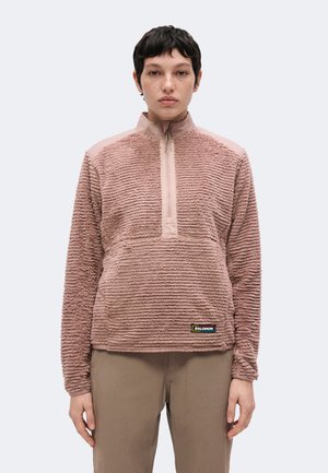 Person med kort mørkt hår iført en tekstureret støvet lyserød fleece med kvart lynlås og lysebrune bukser, set forfra mod en ensfarvet baggrund.