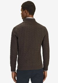 Brun kabelstickad tröja med hög krage, ribbad mudd och nederkant. Har ett strukturerat mönster genom hela plagget, tillverkat av ett mjukt material.