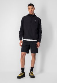 Giacca nera con cappuccio regolabile e tasca frontale, abbinata a pantaloni corti neri e scarpe nere con dettagli gialli e calze nere.