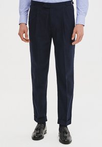 Granatowe spodnie z zakładkami, dopasowany krój i pętle na pasek, w połączeniu z czarnymi butami eleganckimi. Gładka tekstura materiału i wykończenie mankietów w kroju.