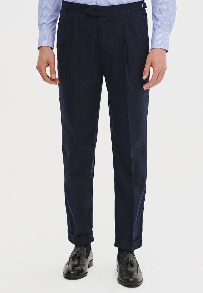 Granatowe spodnie z zakładkami, dopasowany krój i pętle na pasek, w połączeniu z czarnymi butami eleganckimi. Gładka tekstura materiału i wykończenie mankietów w kroju.
