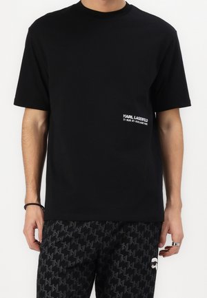 Mężczyzna w czarnym t-shircie z krótkim rękawem z białym napisem Karl Lagerfeld oraz wzorzystych czarnych spodniach z małym logo kreskówki na lewej nodze.