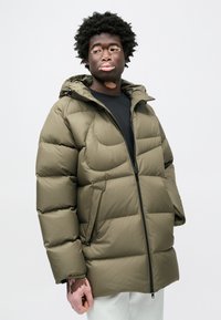 Olivno zelena puhasta jakna z zadrgo spredaj, velikimi žepi in kapuco. Ima prešito obliko in mehko, gladko teksturo.