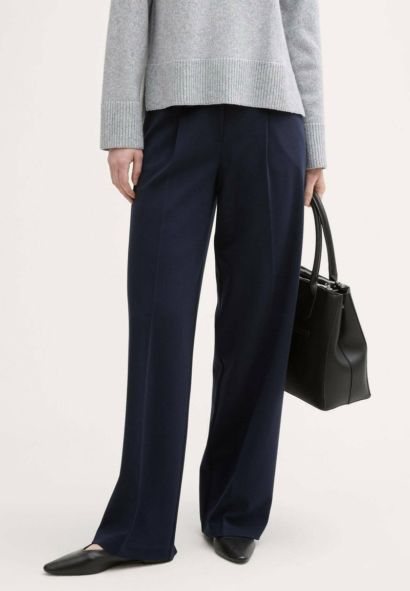 Pantalons larges bleu marine, associés à un pull en tricot gris clair. Un sac à main en cuir noir et des ballerines à bout pointu complètent le look.