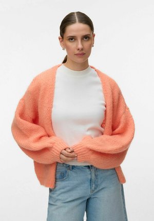 Ung kvinne med brunt hår samlet bak, iført perleøreringer, hvit topp, oversized ferskenfarget strikkegenser og lyseblå jeans mot en hvit bakgrunn.