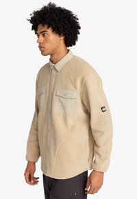 Giacca in pile beige con chiusura a bottoni, due tasche sul petto e colletto a contrasto. Presenta una toppa con logo sulla manica.