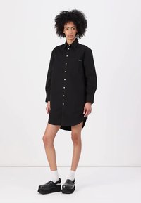Abito camicia nero con bottoni, realizzato in cotone, con maniche lunghe, colletto e spacco laterale; presenta bottoni bianchi e una tasca sul petto.