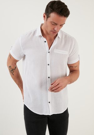 Camisa blanca de manga corta con cierre de botones en la parte delantera, botones negros, un bolsillo en el pecho y una tela texturizada. Combinada con pantalones negros.