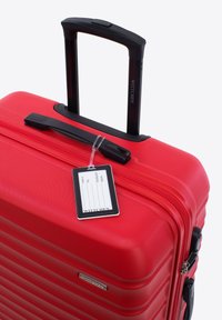 Maleta rígida roja con superficie texturizada, asa extensible individual y etiqueta de equipaje adjunta. Cuenta con un cierre de cremallera y el logo de la marca.