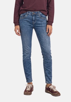 Personne portant un jean slim bleu, un sweat bordeaux et des baskets bordeaux, debout devant un fond clair uni.