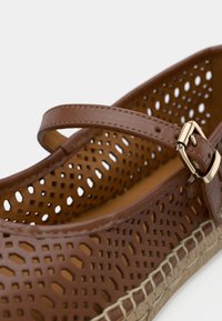 Καφέ τρυπητό δερμάτινο παπούτσι με λουράκι και χρυσή πόρπη. Διαθέτει υφασμένη σόλα εσπαντρίγιας, που ενισχύει την αναπνευσιμότητα και το σχέδιο.