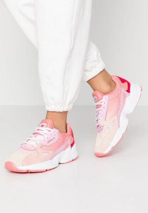 Voeten met roze en witte Adidas sneakers gecombineerd met witte joggerbroeken tegen een effen lichtgrijze achtergrond.