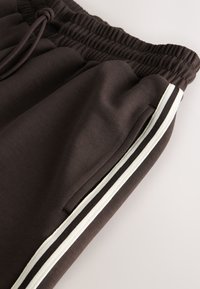 Braune Jogginghose mit elastischem Bund und Kordelzug sowie Seitentaschen, verziert mit weißen und schwarzen Streifen an den Seiten.