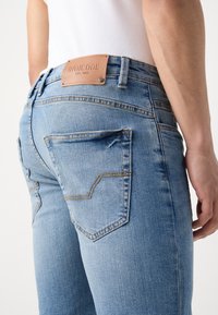 Jean en denim bleu clair avec un patch de marque en cuir sur la ceinture et une poche arrière visible, porté avec un haut blanc.