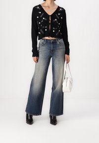 Cárdigan cortado negro con bordado floral, combinado con unos vaqueros de pierna ancha en azul claro. La modelo sostiene un pequeño bolso blanco y lleva botas negras.